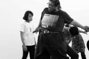 A male performer grimaces, holding hands with another performer bending low while a woman looks on.