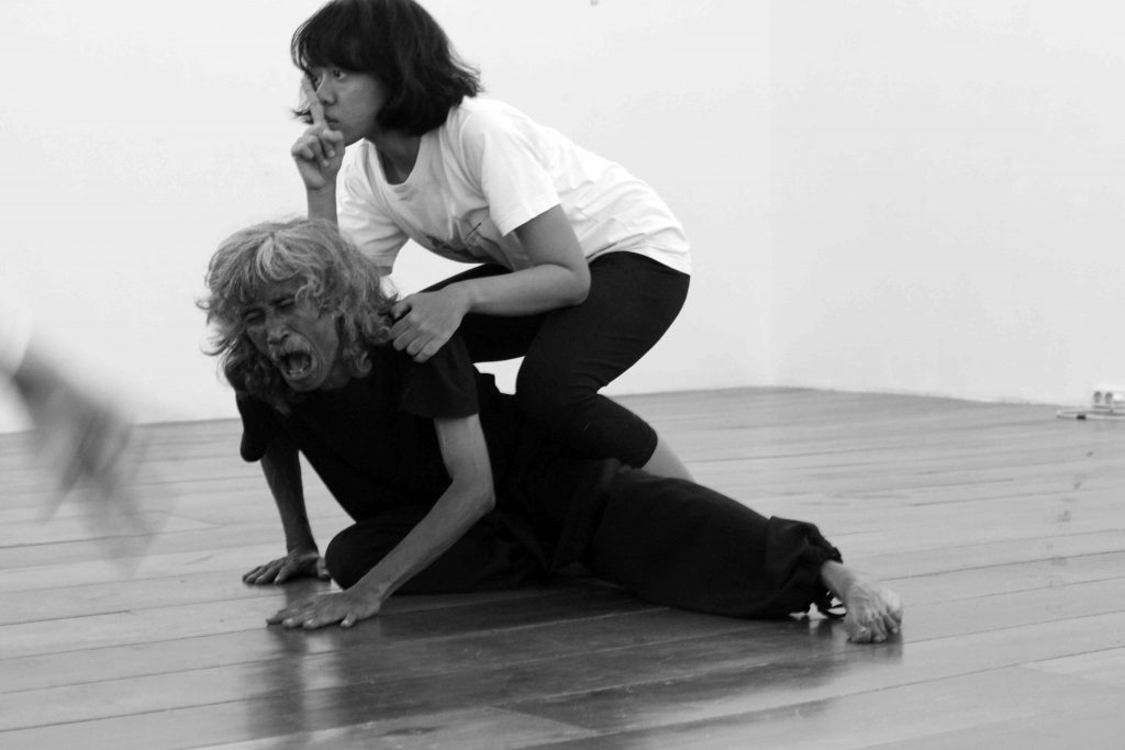 A woman performer with forefinger held up for silence kneels on the back of a scowling male performer.