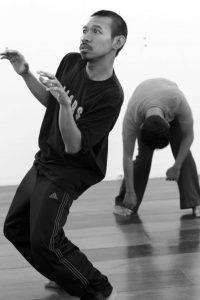 An actor with frightened expression leans to one side in front of another actor bent over from the waist