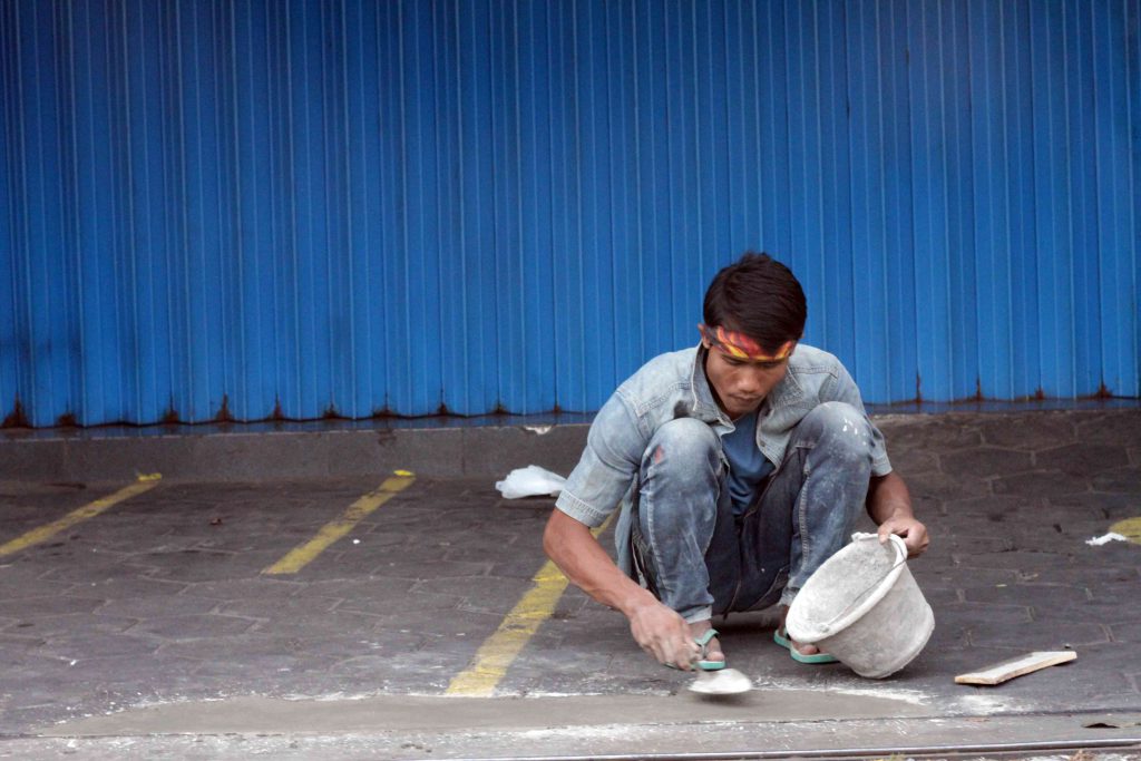 A man squats on the street repairing the sidewalk with a bucket of cement and trowel.