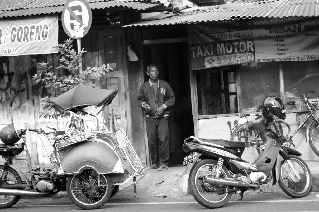 Photo shows a man standing in the doorway of a taxi stand beside two motorbikes.