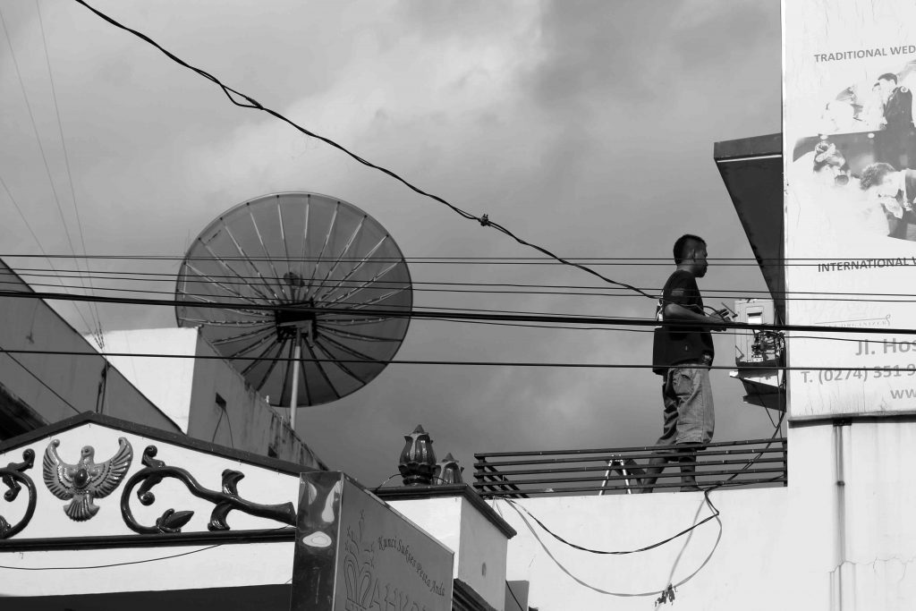 Cityscape shows a man standing on a roof beside a satellite dish.