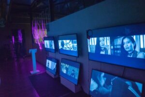 Colour photo shows installation view of video exhibition with multiple screens showing split screen images of men and women.