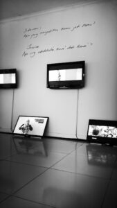 Exhibition view of video installation with monitors hung on wall and resting on the floor, beneath words handwritten on the wall.