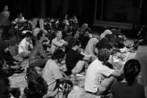 A group of people sits on the floor, facing forward and listening.
