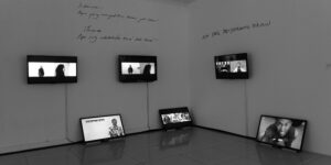 Exhibition view of video installation with monitors hung on walls and resting on the floor, as well as words written on the wall.